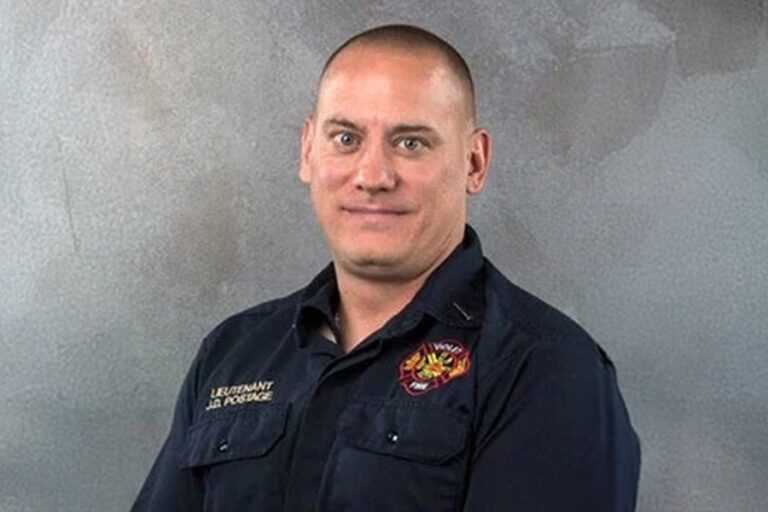 A person in a dark uniform with an embroidered badge and text on the chest stands against a gray background.