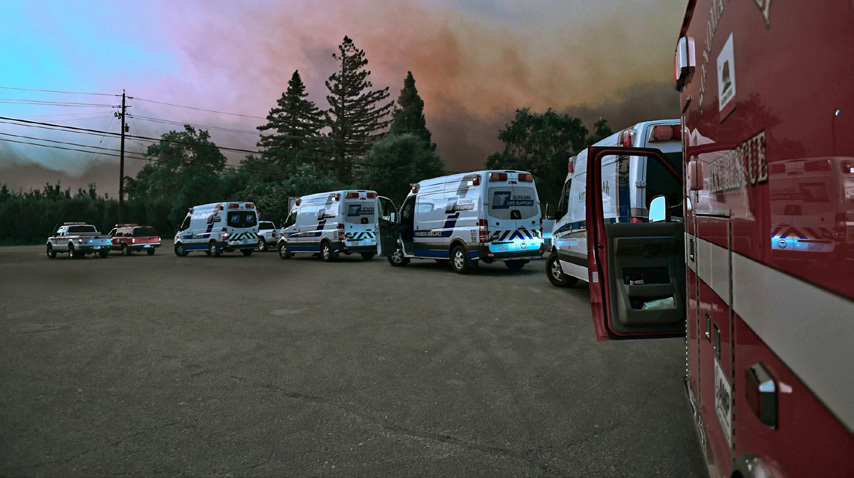 Line of emergency vehicles on a road, with several ambulances and police cars under a smoky sky, indicating an emergency situation.