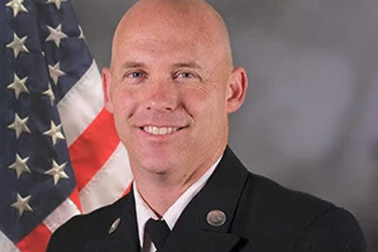 Smiling person in a dark suit stands in front of an American flag backdrop.