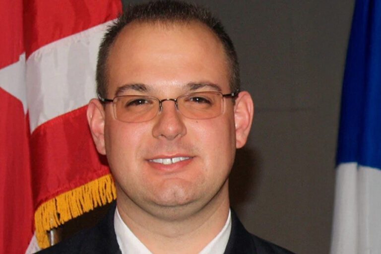 A man wearing glasses, a suit, and a tie stands in front of a red and white flag.
