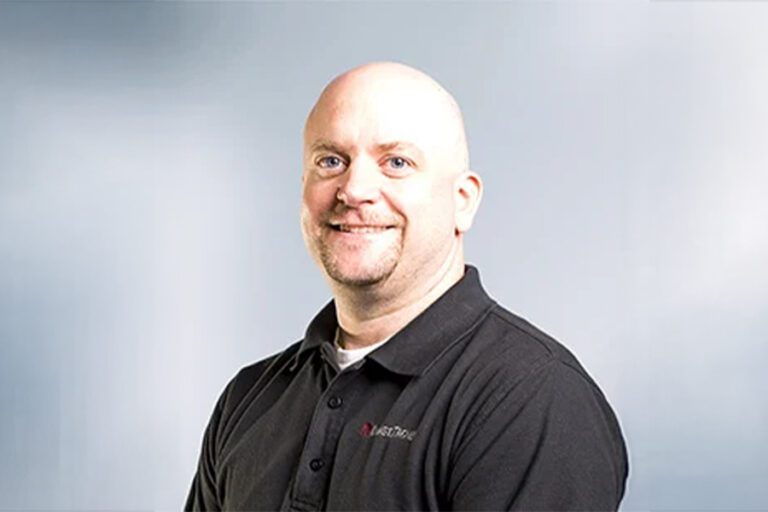 A bald man in a black polo shirt smiles while standing in front of a light gray background.