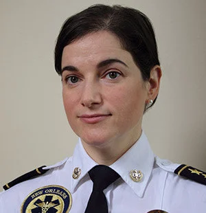 Person in New Orleans emergency services uniform, wearing a white shirt and black tie, looks towards the camera against a plain background.