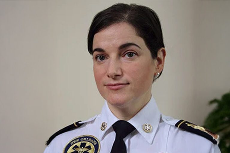 Person in a white police uniform looking straight ahead, with badge details and a plant partially visible in the background.