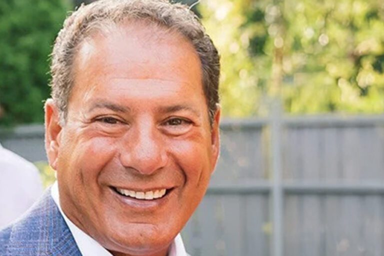 Smiling man in a blue suit jacket stands outdoors with a blurred green and gray background.