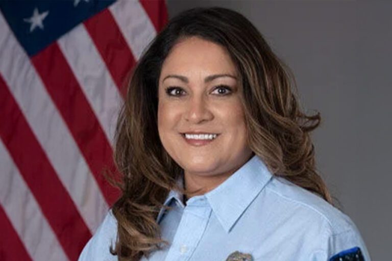 A person in a light blue uniform shirt smiles in front of a U.S. flag backdrop.