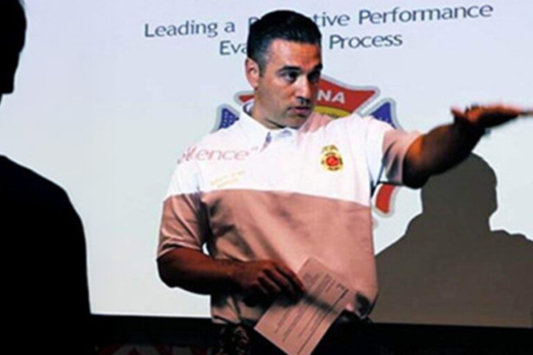 A man in a white polo shirt gestures in front of a presentation slide. He holds a paper in his other hand.