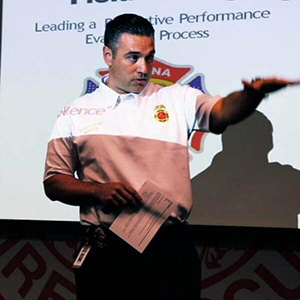A person in a polo shirt gives a presentation, gesturing with one hand. Text and a logo are projected in the background.