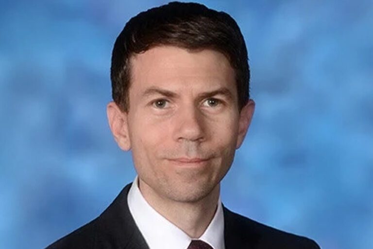 A man in a dark suit and tie with short brown hair poses against a blue background.