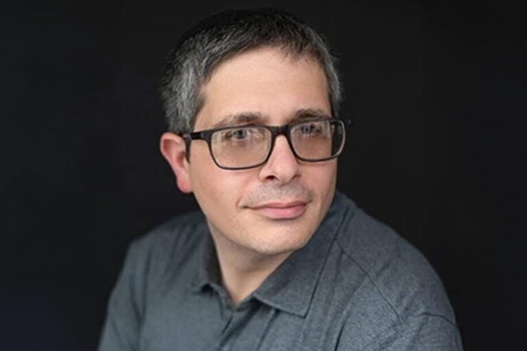 Man with glasses, wearing a grey shirt, looking off to the side against a dark background.