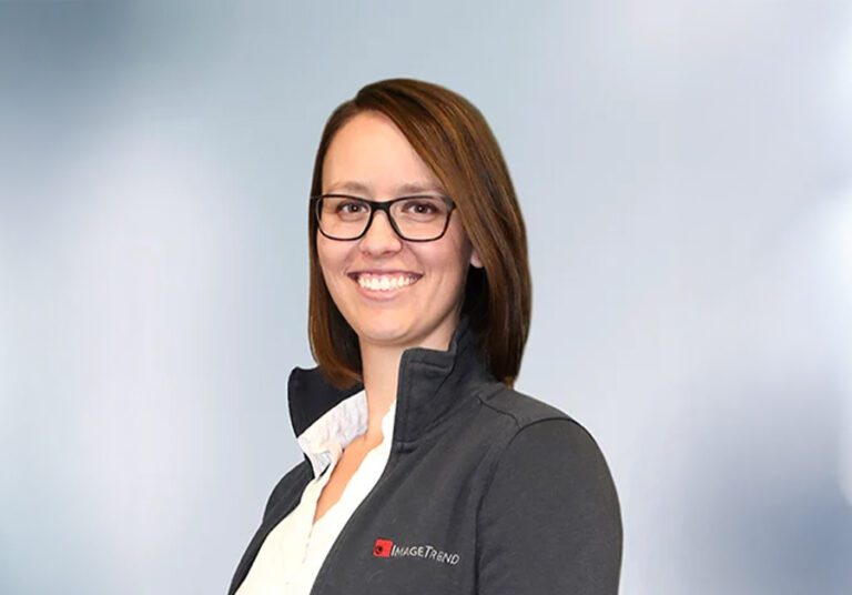 Smiling person with brown hair and glasses wearing a dark jacket over a white shirt, against a light gray background.