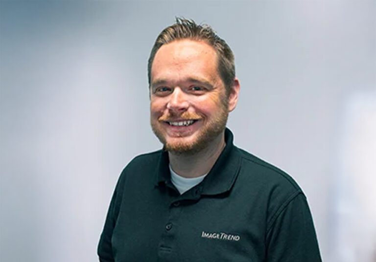 Man with short hair and beard smiling, wearing a dark polo shirt with a logo, stands against a plain light background.
