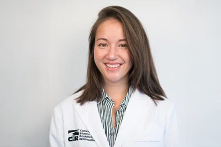 Smiling person with shoulder-length hair wearing a white lab coat over a striped shirt.