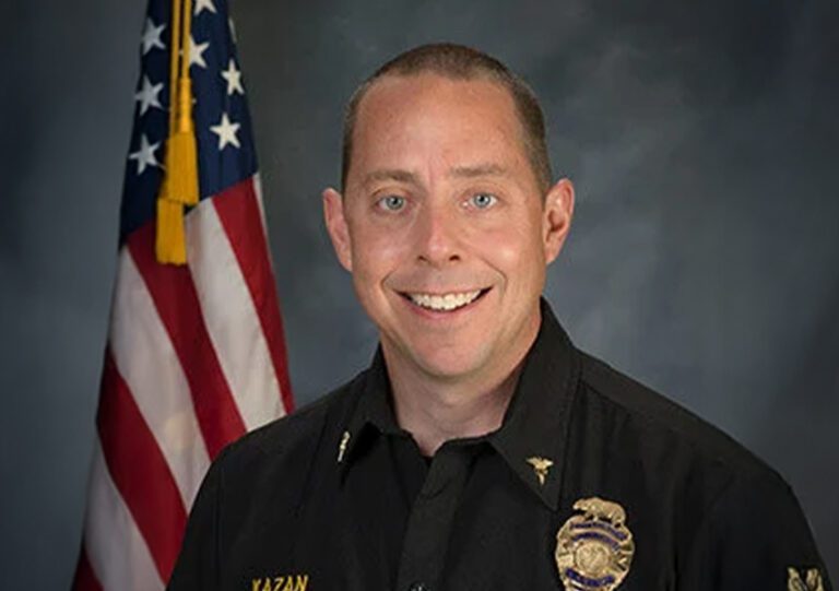 Smiling individual in a dark uniform stands in front of an American flag.