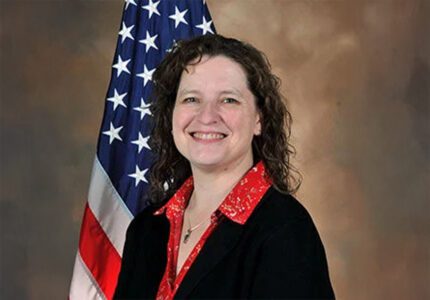 A person with curly hair smiles while standing in front of the U.S. flag. They are wearing a black blazer over a red shirt.