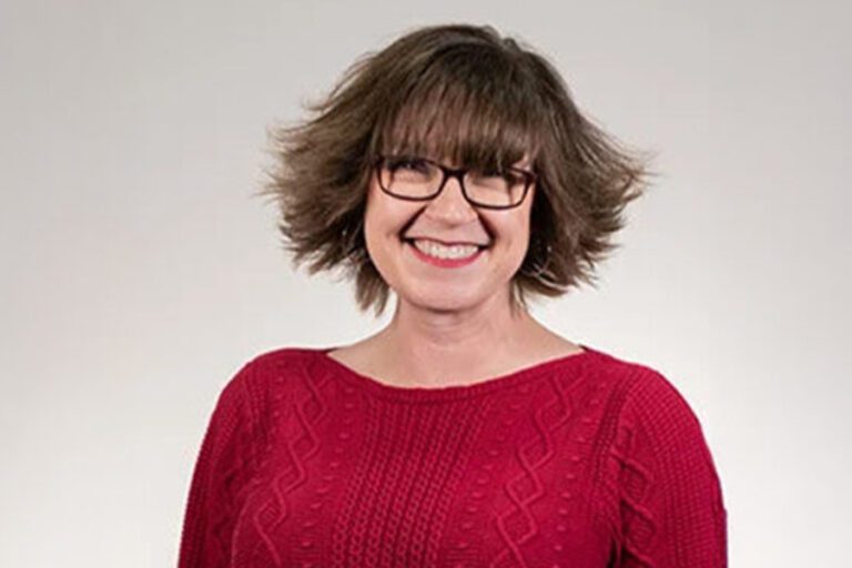 Person with short brown hair, glasses, and a red sweater smiles against a plain background.
