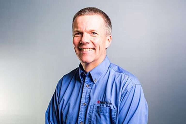 A person wearing a blue button-up shirt with embroidered text, smiling against a plain background.