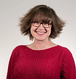 Woman with wavy hair wearing glasses and a red sweater smiles against a plain background.