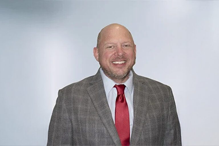 A man in a plaid suit with a red tie smiles against a plain background.