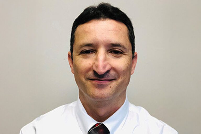 Man in a white shirt and tie against a plain background, smiling slightly at the camera.