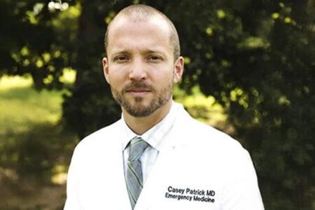 A person with a beard in a white lab coat stands outdoors, with trees in the background. The coat reads, "Casey Patrick MD Emergency Medicine.