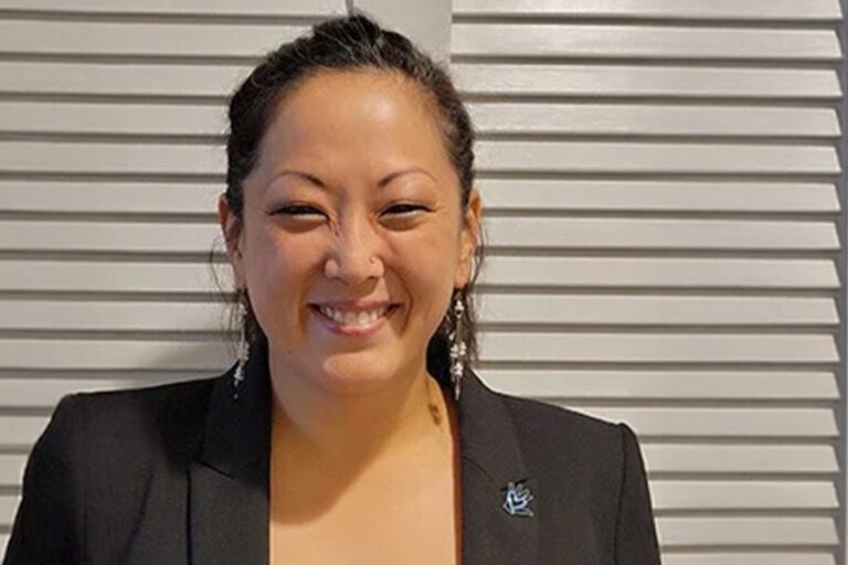 Smiling woman in a black blazer stands in front of a white, slatted wall.