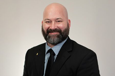 A bald man with a beard wearing a black suit and a checkered shirt smiles in front of a plain background.