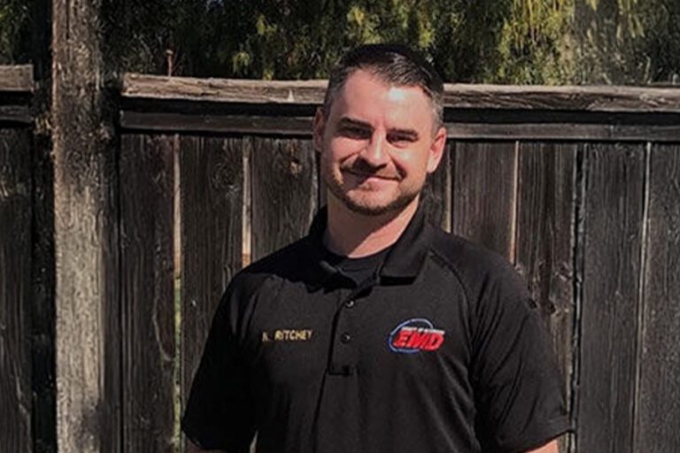 A man in a black polo shirt with embroidered text stands in front of a wooden fence, outdoors.