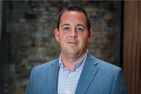 Man in a blue suit jacket and checkered shirt standing in front of a brick wall.