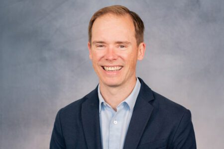 A person with short hair smiles, wearing a blue shirt and dark blazer, in front of a gray background.