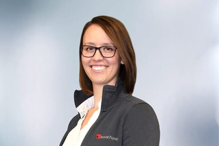 A woman with brown hair and glasses smiles in front of a plain background. She is wearing a dark jacket with a logo on it.