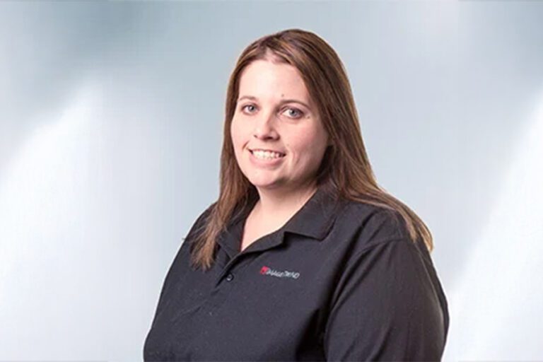 A woman with long brown hair smiles, wearing a black shirt with a red and white logo on the left side, against a light gray background.