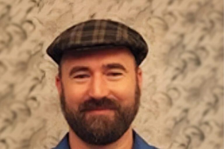 A man with a beard wearing a plaid flat cap and a blue shirt stands against a patterned background.