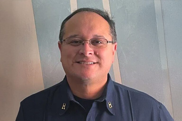 A person wearing glasses and a dark shirt stands in front of a light-colored wall, smiling.