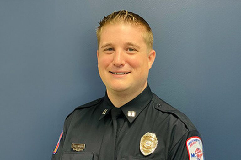A man in a dark police uniform with insignia and badges, standing against a plain blue background, smiling lightly.