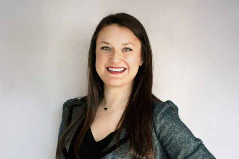 A woman with long brown hair smiles while standing against a plain background, wearing a dark top and a shimmering jacket.