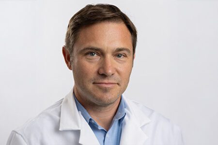 A person wearing a white lab coat and blue shirt against a plain background.