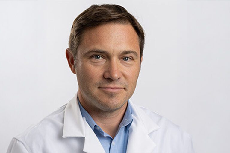A person wearing a white lab coat and blue shirt against a plain background.