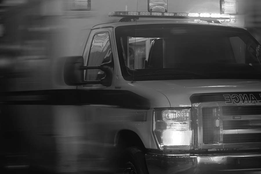 Black and white image of an ambulance with its lights flashing, captured from the front side, partially blurred to indicate motion.