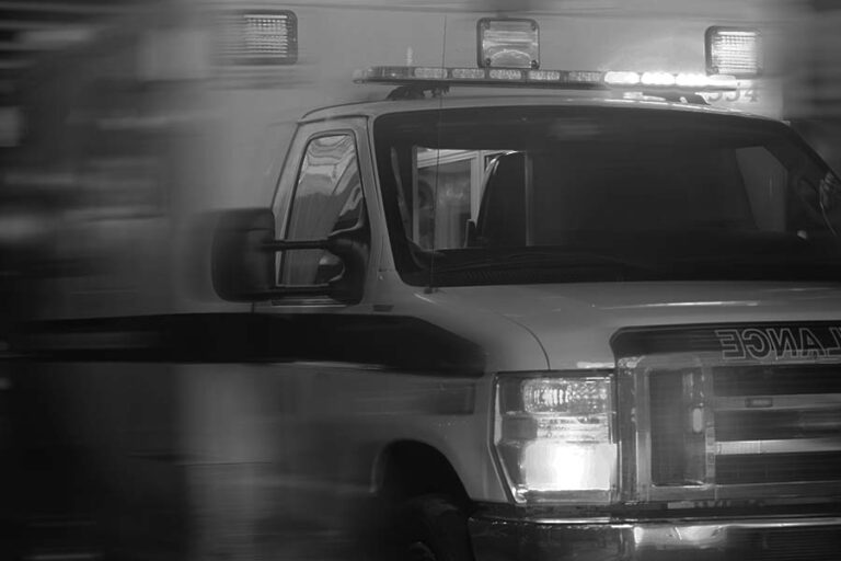 Black and white image of an ambulance with flashing lights in motion, viewed from the side.