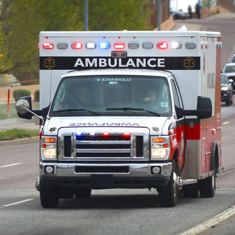 A red and white ambulance drives down a road with flashing lights on, surrounded by other vehicles.