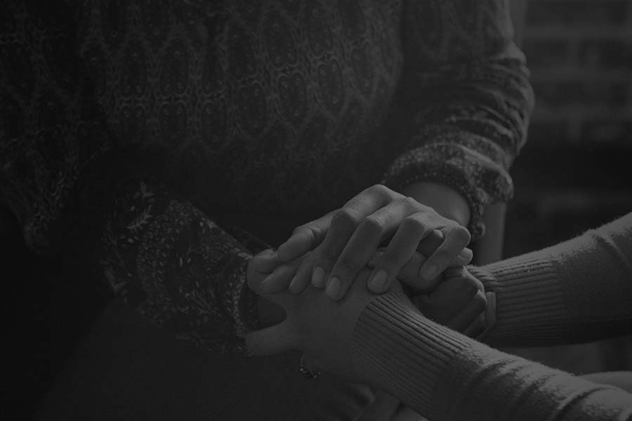 Two people sitting and holding hands, one wearing a patterned sweater and the other in a plain long-sleeve shirt, against a dark blurred background.