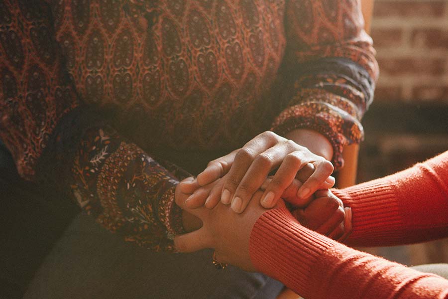 Two people sitting, one wearing a patterned top and the other an orange sweater, holding hands in a gesture of comfort and support.