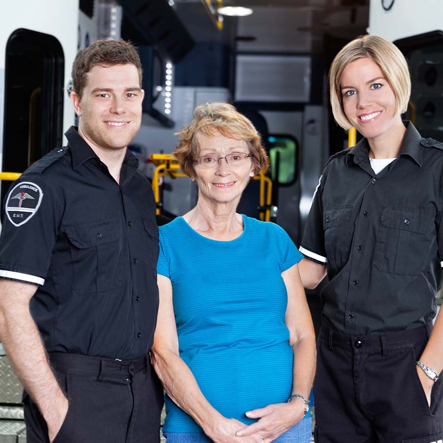 Two paramedics in uniform stand on either side of an older woman, smiling, in front of an ambulance.