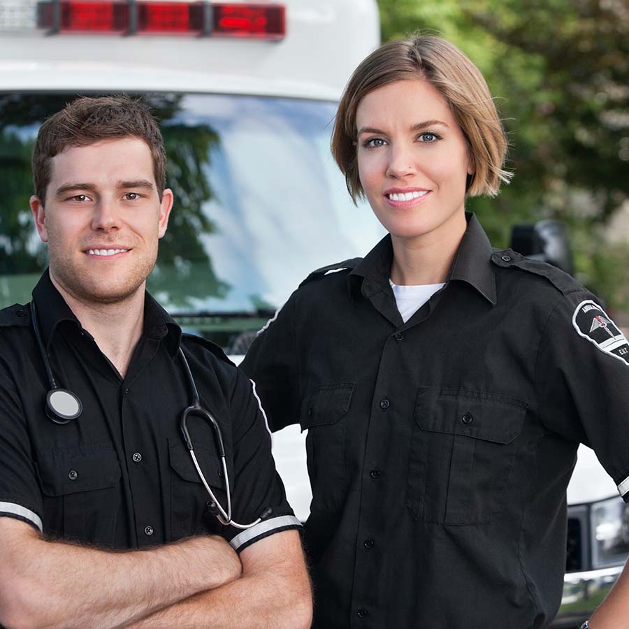 Two paramedics in black uniforms stand in front of an ambulance. One has a stethoscope around the neck. Both are smiling at the camera.