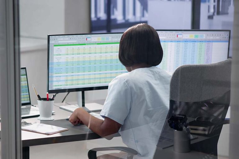 Person in an office setting working on a computer with spreadsheets, surrounded by a clipboard and pen holder.