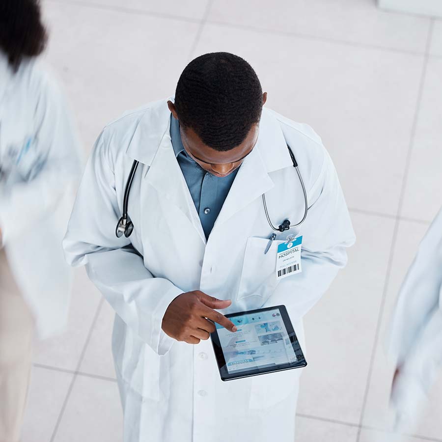 A doctor in a white coat and stethoscope uses a tablet, standing on a light-colored tiled floor.