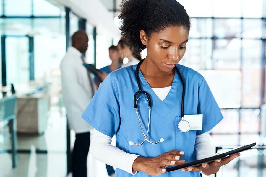 A healthcare professional in blue scrubs and a stethoscope uses a tablet in a modern hospital setting as colleagues converse in the background.