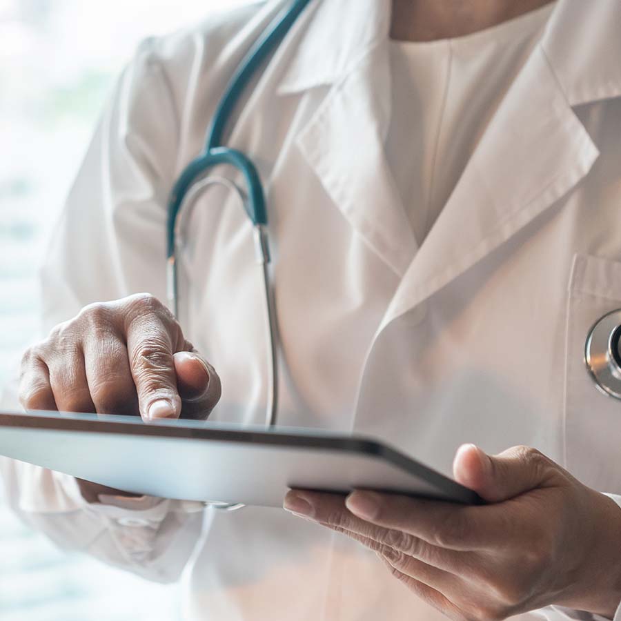 A doctor in a white coat uses a tablet, with a stethoscope around their neck.