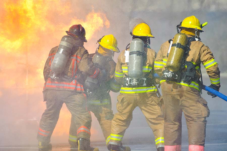 Four firefighters in gear and helmets, with oxygen tanks, tackle a blaze with a hose. Flames and smoke are visible in the background.