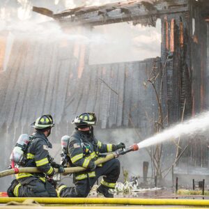 two firefighters kneeling and holding hose as it sprays water on fire